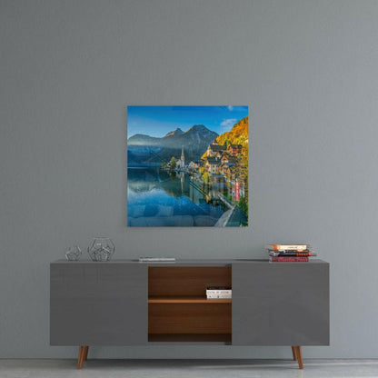 Square glass wall art depicting Alpine lake village with church spire, mountains, and autumn foliage, displayed above modern console table.