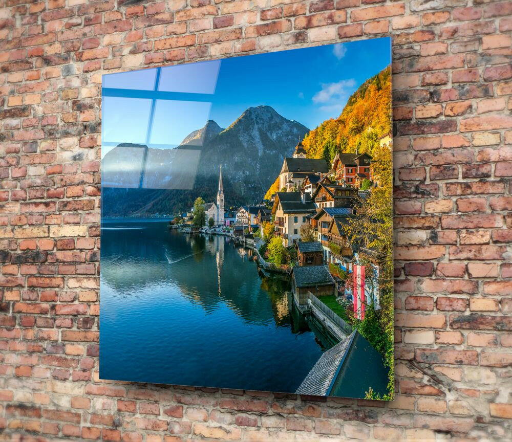 Glass wall art displaying Hallstatt village with alpine lake, mountains, and autumn foliage mounted on red brick wall.