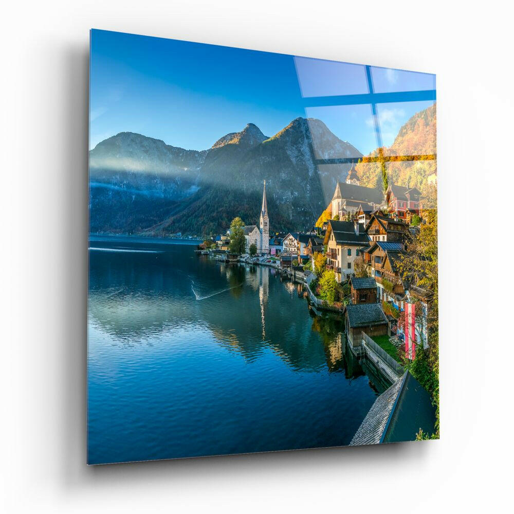 Glass wall art mounted on white wall displaying Alpine lakeside village with church, colorful houses, mountains, and blue water.
