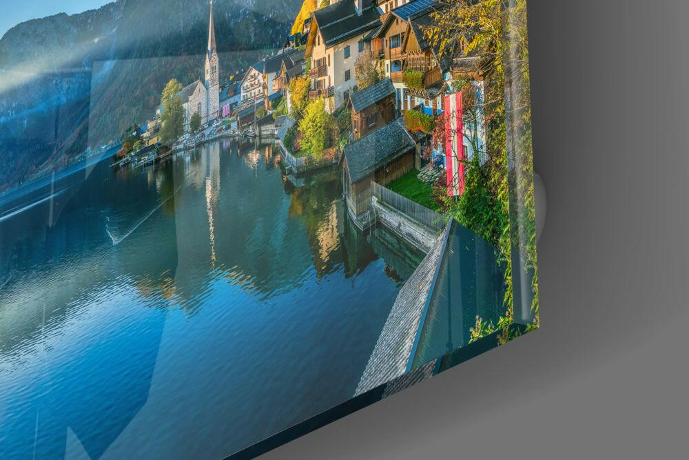 Angled perspective of glass wall art showing Alpine lakeside village with church steeple, colorful buildings, and blue water reflections.