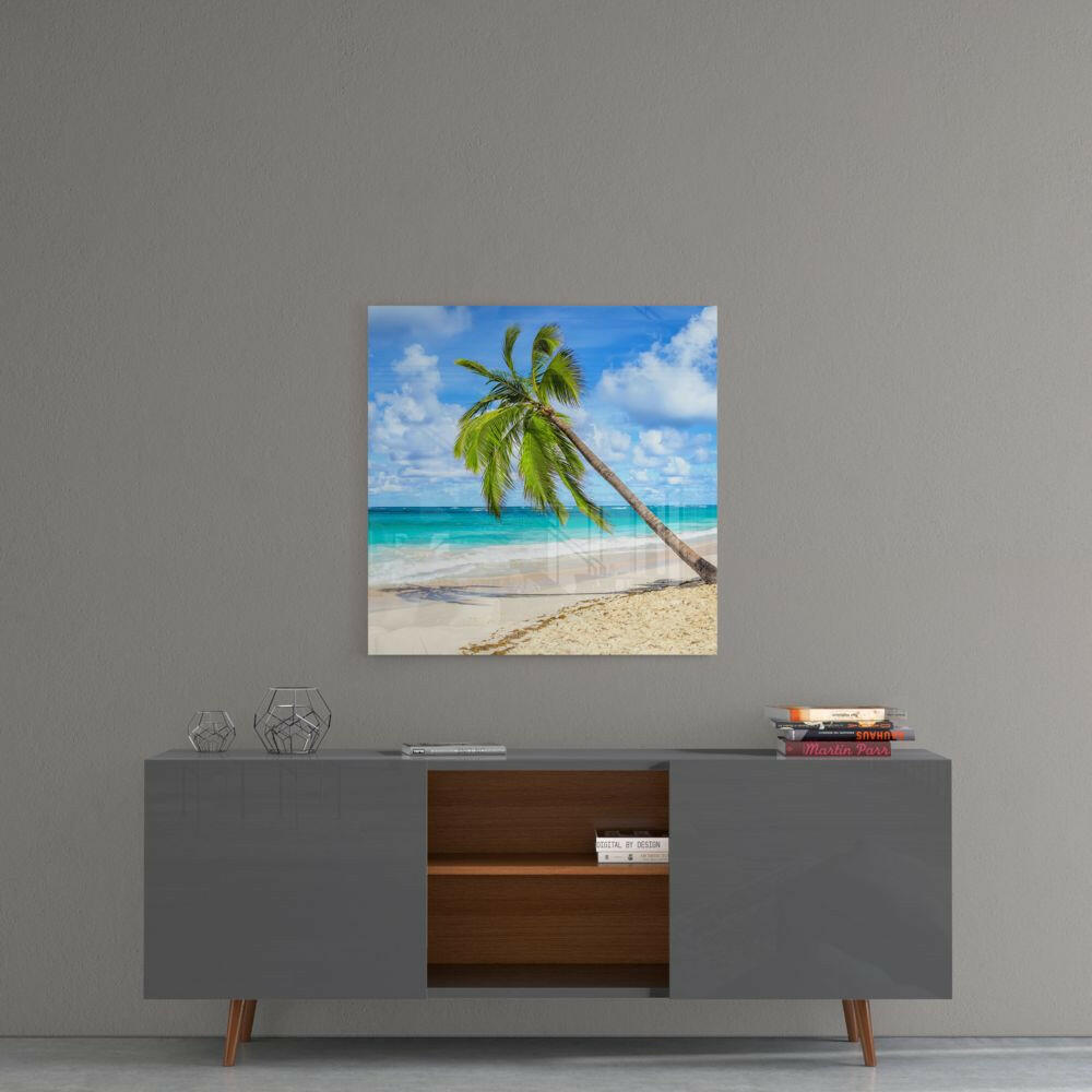 Square glass wall art depicting a leaning palm tree on a sandy beach with turquoise ocean and blue sky, displayed above a gray mid-century modern dresser.