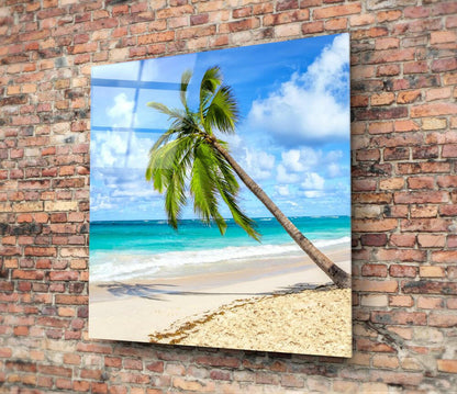 Glass wall art mounted on red brick wall displaying tropical beach scene with leaning palm tree, white sand, turquoise ocean, and blue sky.