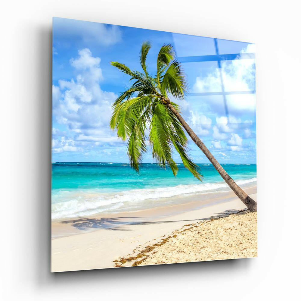 Angled view of glass wall art displaying a tropical beach scene with a leaning palm tree, turquoise ocean, and blue sky.