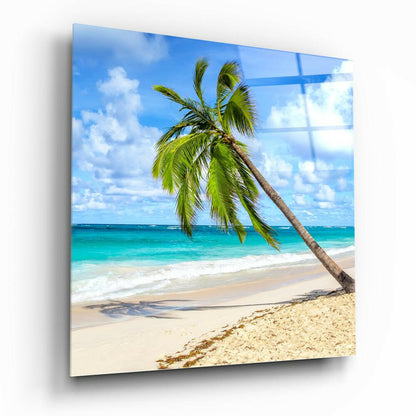 Angled view of glass wall art displaying a tropical beach scene with a leaning palm tree, turquoise ocean, and blue sky.