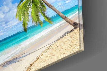 Angled view of glass wall art displaying tropical beach scene with palm frond, turquoise water, and white sand against gray wall.