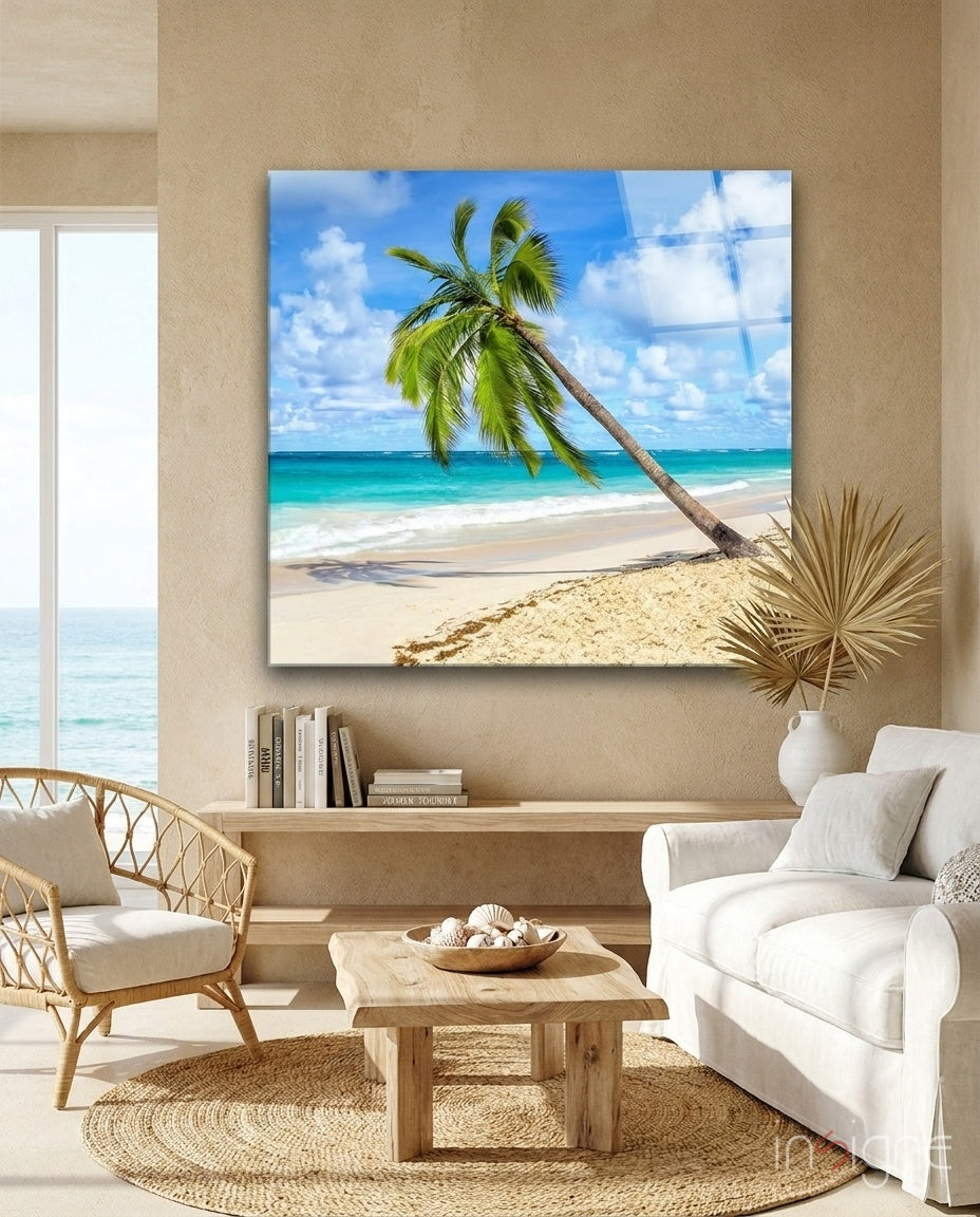 Glass wall art displaying a tropical beach scene with a leaning palm tree, turquoise ocean, and sandy shore hung in a coastal living room.