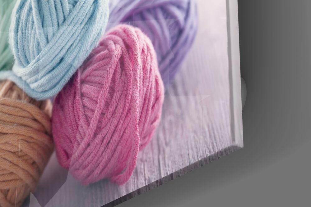 Close-up edge view of glass wall art displaying colorful yarn balls in blue, pink, purple, and tan against white background.