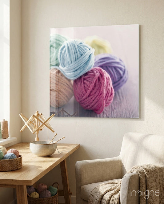 Glass wall art displaying colorful yarn balls in blue, pink, and purple hung above a wooden craft table in a sunlit room.