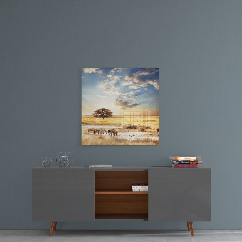 Glass wall art depicting African savanna landscape with acacia tree, elephants, golden grassland, and dramatic sky displayed above gray sideboard.