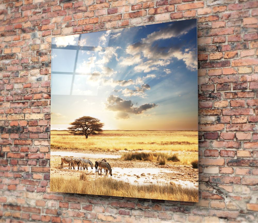 Glass wall art mounted on red brick showing African savanna landscape with zebras at watering hole, acacia tree, golden grassland, and blue sky with clouds.