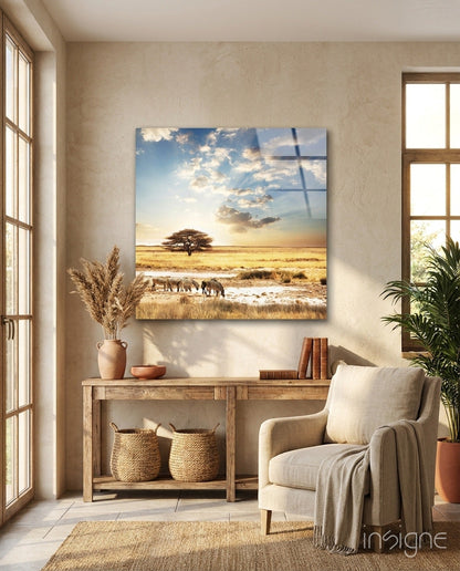 Glass wall art depicting African savanna landscape with acacia tree, elephants, and sunset sky, displayed above wooden console table in light room.