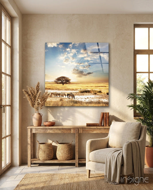 Glass wall art depicting African savanna landscape with acacia tree, elephants, and sunset sky, displayed above wooden console table in light room.