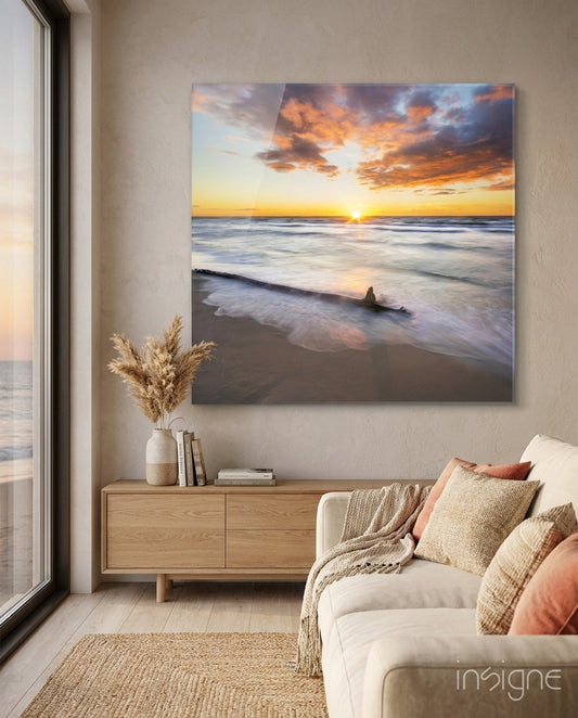 Sunset beach scene with golden sky and waves displayed as glass wall art above wooden cabinet in coastal living room.