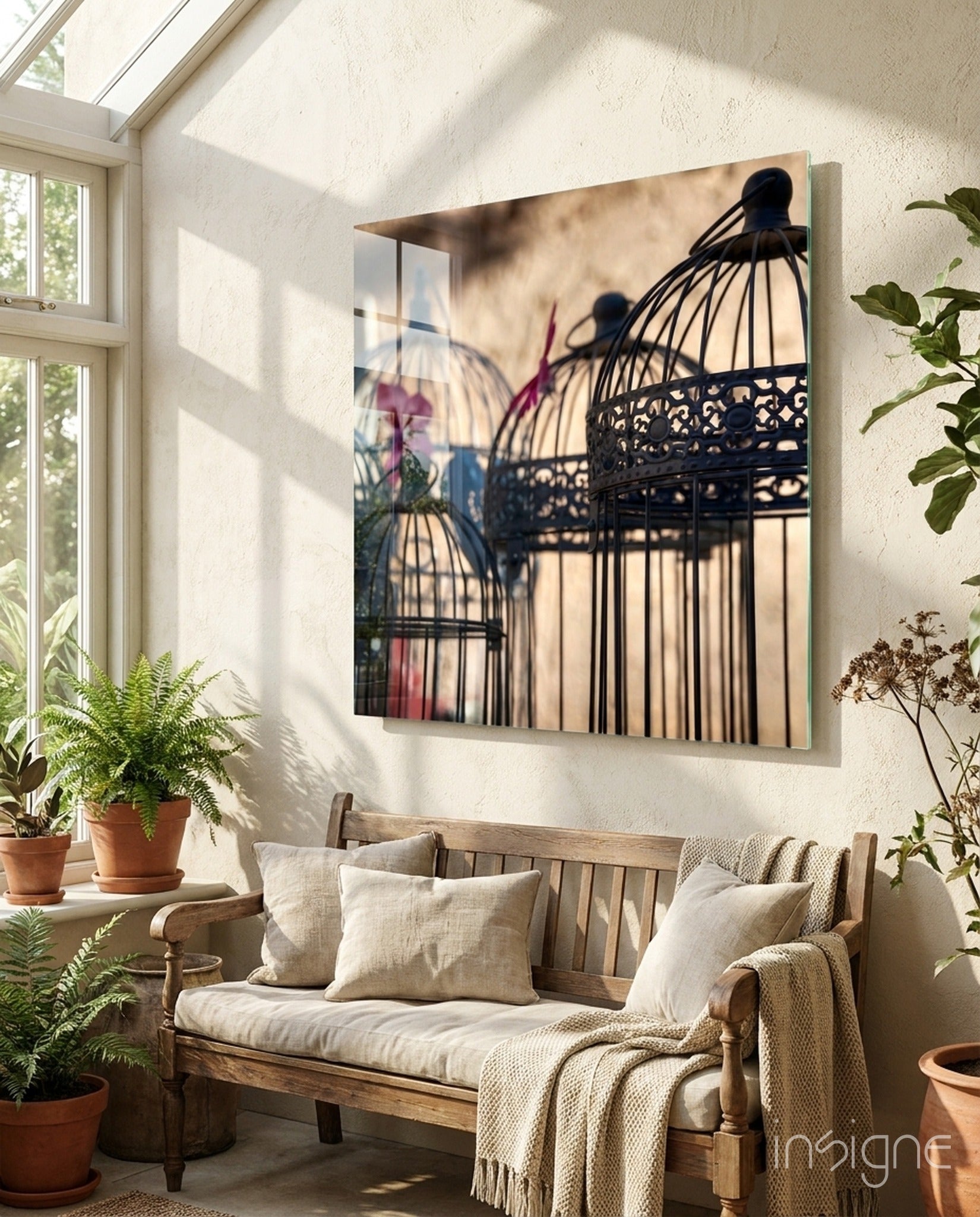 Glass wall art depicting ornate black birdcages with colorful fabric shadows displayed above a wooden bench in sunlit room.