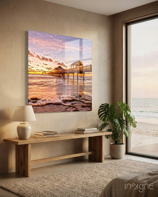 Pink Sunset Pavilion Dock glass wall art displayed above wooden console table in coastal bedroom interior with ocean view window.