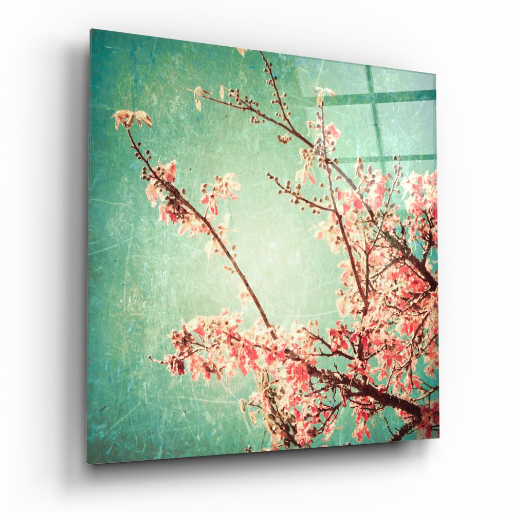 Angled view of glass wall art featuring coral-pink flowering branches against a turquoise-green textured background with grid pattern.