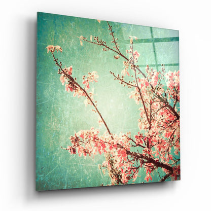 Angled view of glass wall art featuring coral-pink flowering branches against a turquoise-green textured background with grid pattern.