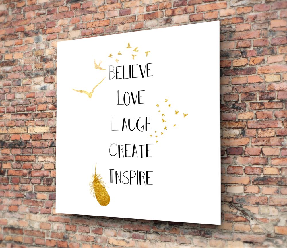 White glass wall art with gold foil birds and text reading "Believe Love Laugh Create Inspire" mounted on red brick wall.