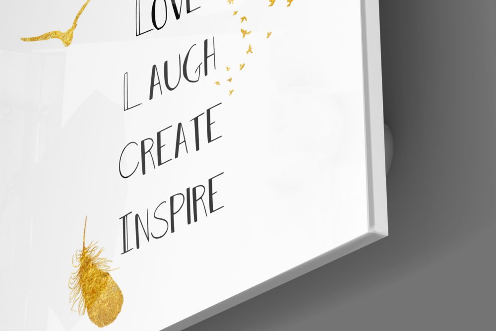 Angled view of glass wall art with black handwritten text reading "LOVE LAUGH CREATE INSPIRE" and gold decorative leaf elements on white background.