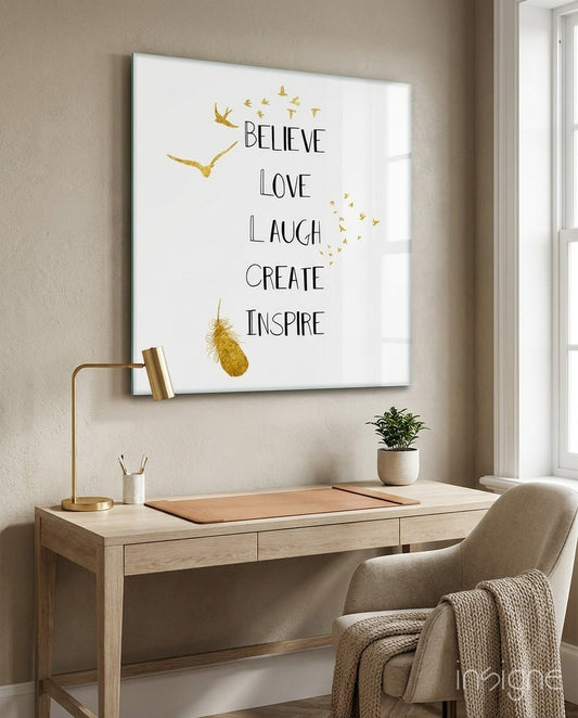 Glass wall art with gold birds and inspirational text "Believe Love Laugh Create Inspire" displayed above a wooden desk in a minimalist home office.