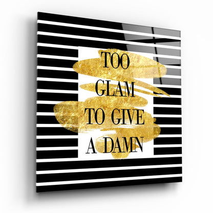 Angled view of glass wall art with black and white horizontal stripes, gold brushstroke shapes, and text reading "TOO GLAM TO GIVE A DAMN.