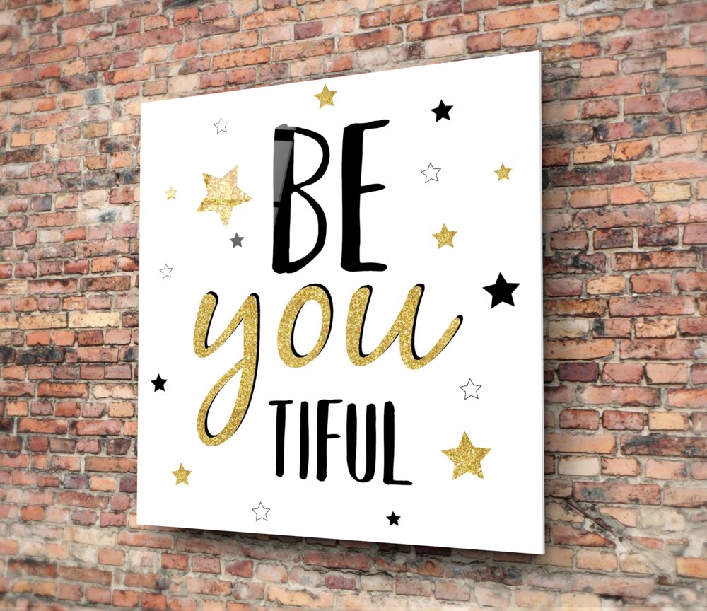White glass wall art with "BE you TIFUL" text in black and gold lettering, surrounded by gold and black stars, mounted on red brick wall.