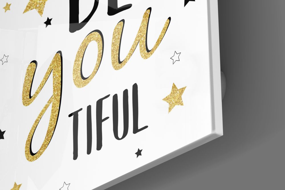 Close-up angled view of white glass wall art with gold glitter script reading "You Are" and black text "TIFUL" with scattered gold and black stars.