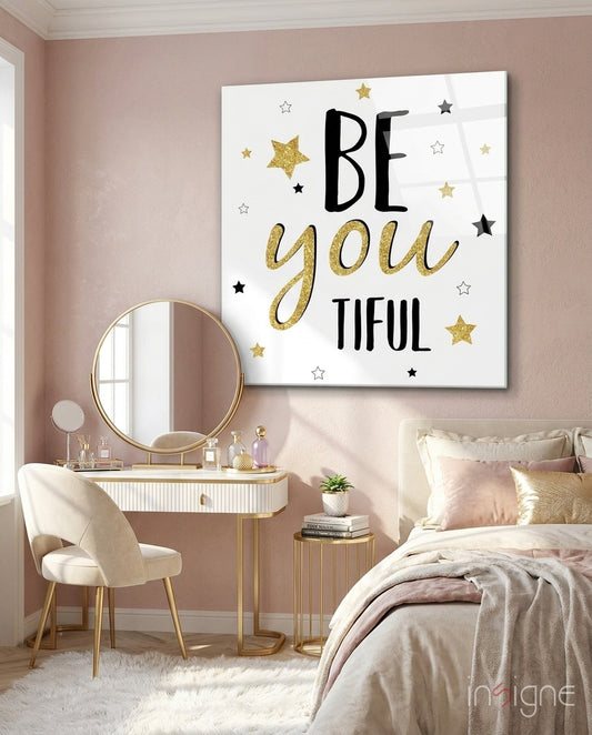 Motivational glass wall art with "BE you TIFUL" text and gold and black stars displayed above a vanity in a pink bedroom.