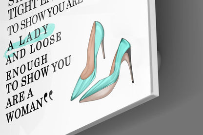 Angled view of glass wall art displaying turquoise high heels with empowerment text on white background.