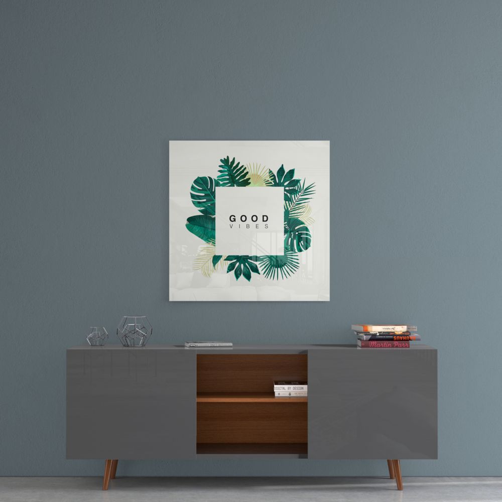 Square glass wall art with "GOOD VIBES" text surrounded by green and gold botanical leaf illustrations, displayed above a modern credenza.