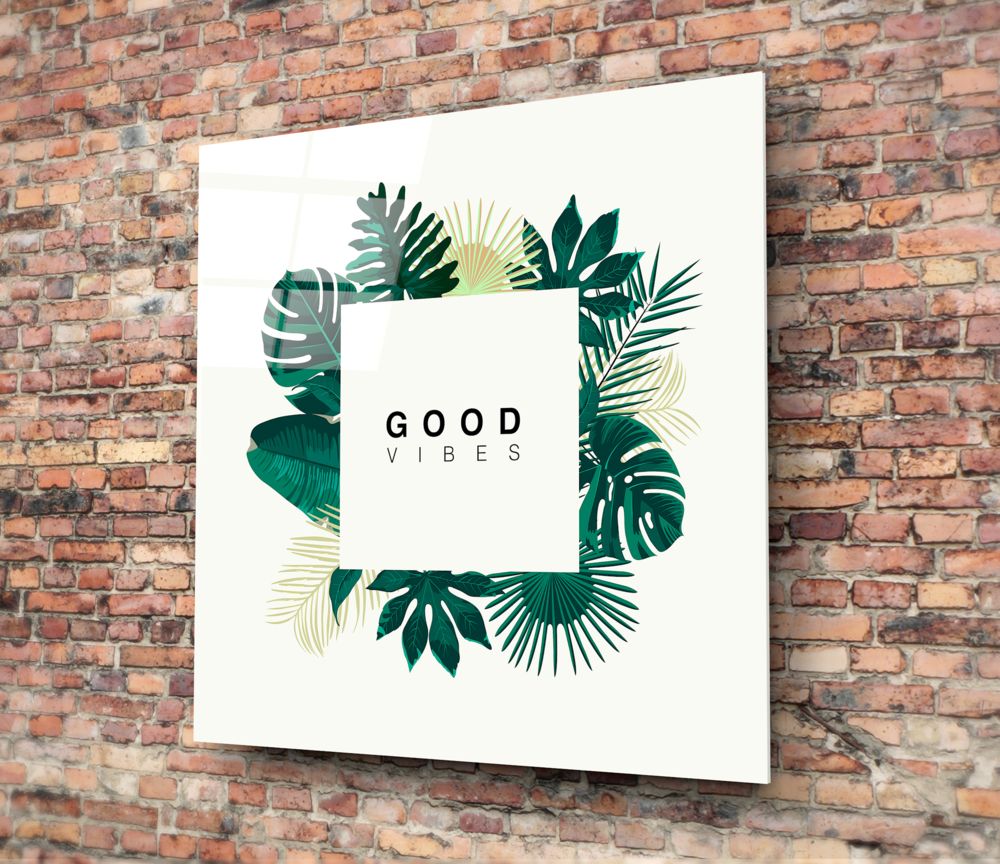 White glass wall art with "GOOD VIBES" text surrounded by green and cream tropical leaf illustrations, mounted on red brick wall.