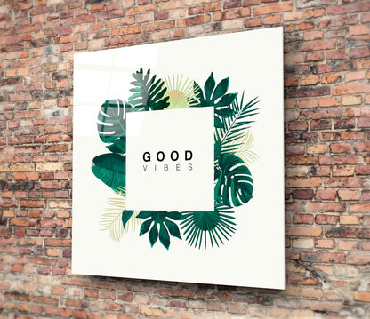 White glass wall art with "GOOD VIBES" text surrounded by green and cream tropical leaf illustrations, mounted on red brick wall.