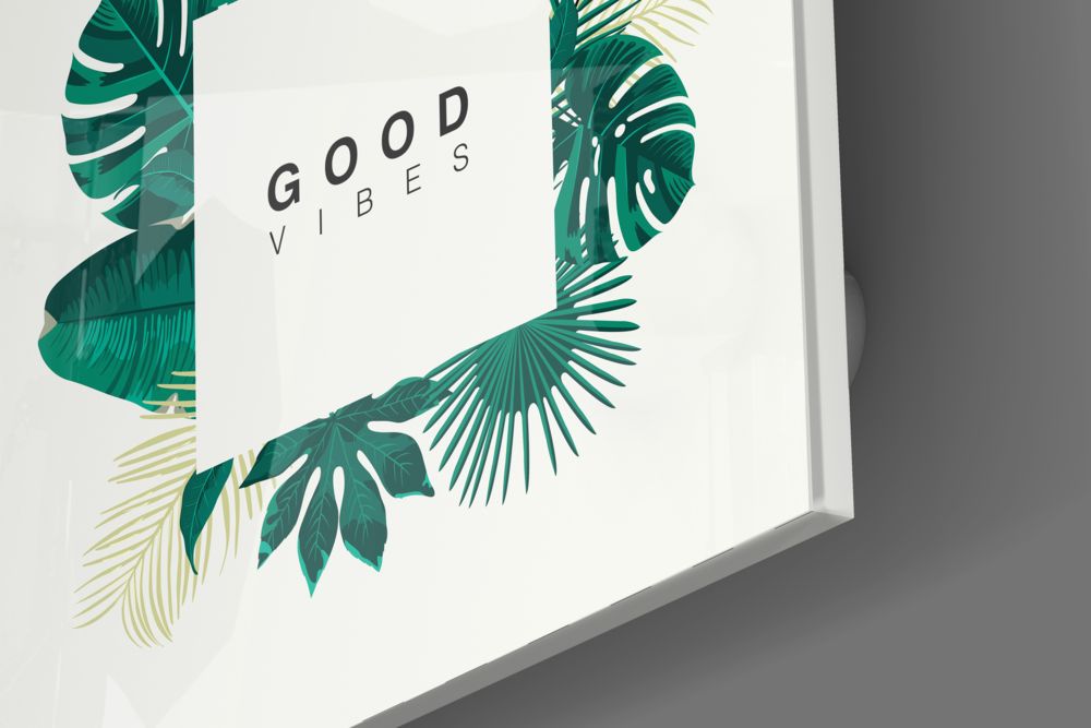 Angled view of white glass wall art with green and gold tropical leaf illustrations and "GOOD VIBES" text centered.
