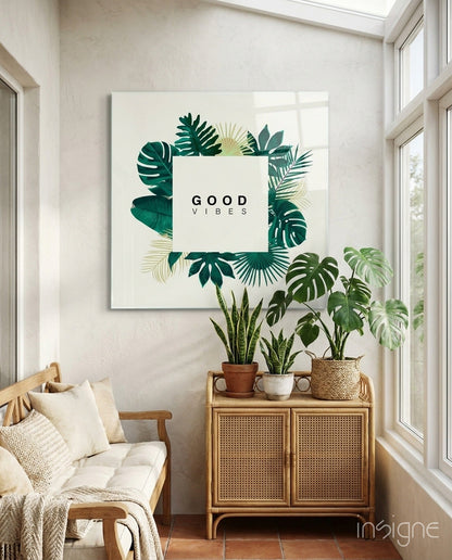 Good Vibes" glass wall art with green tropical leaf illustrations displayed above potted plants in bright sunlit room.