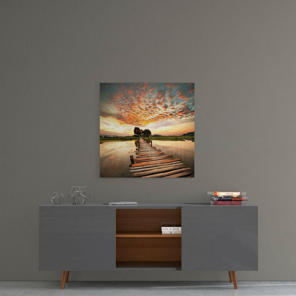 Glass wall art depicting a wooden dock extending into still water at sunset with orange clouds and distant trees.
