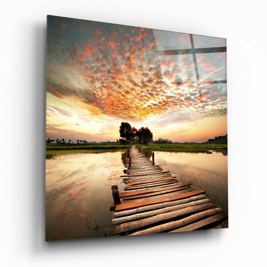 Wooden dock extending into calm water under dramatic orange and golden sunset sky with green fields and trees along horizon.