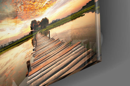 Angled perspective view of glass wall art displaying wooden dock extending toward sunset landscape with golden sky and green fields.