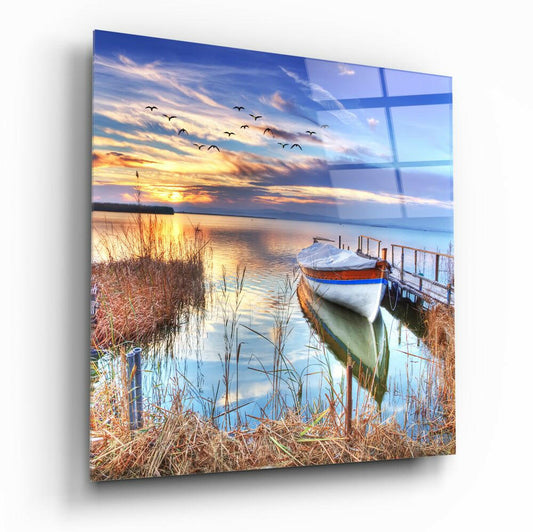 Angled perspective view of glass wall art depicting a moored red and white boat at sunset with reeds, water, and flying birds.