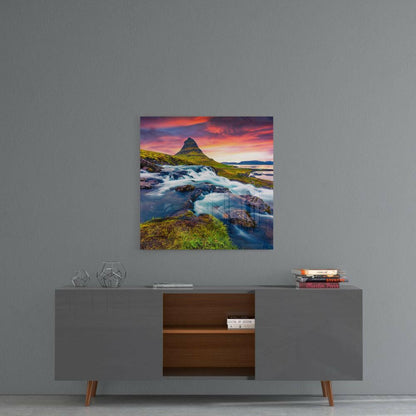 Glass wall art displaying an Icelandic mountain landscape with waterfall, green moss, flowing river, and dramatic sunset sky in orange, pink, and purple tones, hung above a mid-century modern console table.