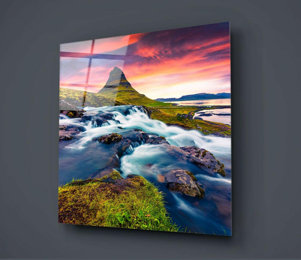 Glass wall art displaying a scenic Icelandic landscape with cascading waterfall, moss-covered rocks, volcanic mountain peak, and dramatic sunset sky in vibrant orange and pink tones.