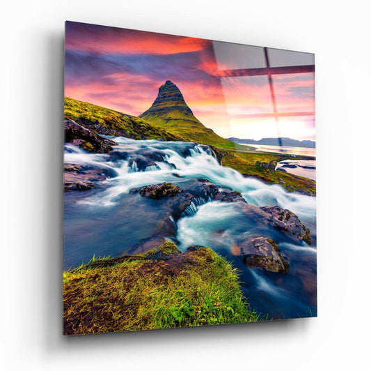 Glass wall art displaying an Icelandic landscape with a cone-shaped mountain, cascading waterfall, moss-covered terrain, and dramatic sunset sky.