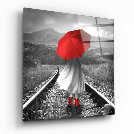 Girl with red umbrella and boots standing on railway tracks facing mountain landscape, black and white with red color accent, glass panel visible.