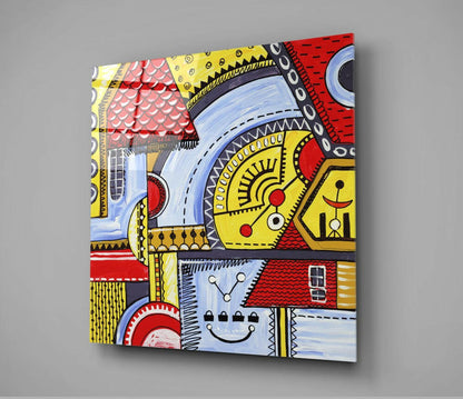 Angled perspective view of a glass wall art panel displaying colorful abstract houses with red roofs, yellow walls, blue windows, and intricate geometric patterns in black outlines.