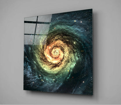 Glass wall art panel displaying a spiral galaxy with glowing yellow center and teal-green outer regions against a starfield, mounted at an angle.