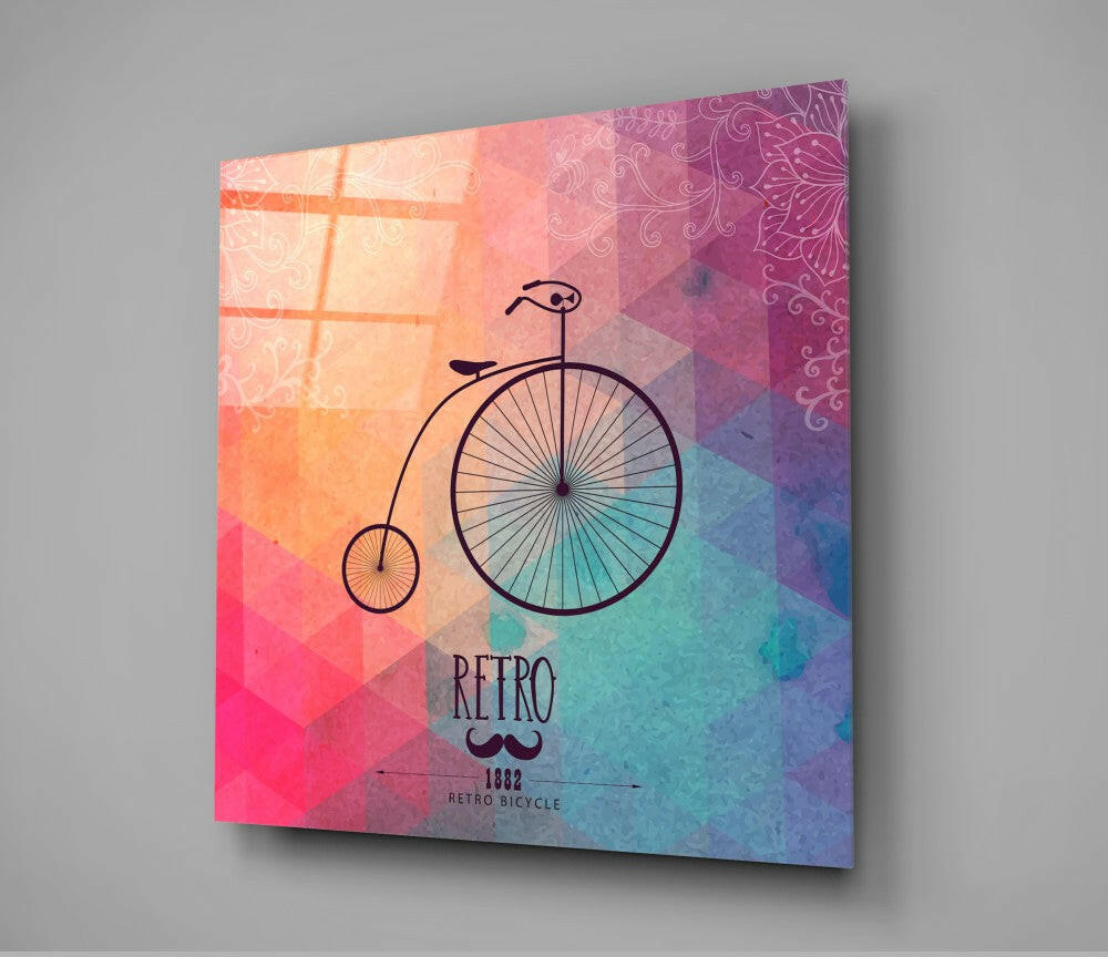 Angled view of glass wall art displaying a penny-farthing bicycle illustration on a gradient background transitioning from coral pink to turquoise with ornamental patterns and "RETRO" text.