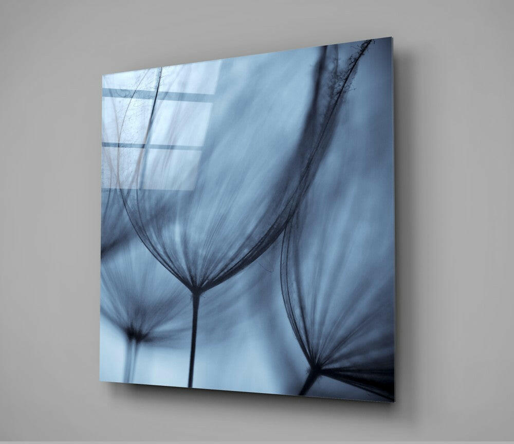 Angled view of glass wall art displaying translucent dandelion seed heads in blue and black tones against light background.