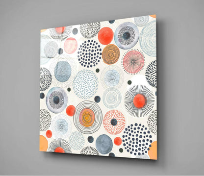 Square glass wall art panel displayed at an angle showing a cream background with colorful geometric circles, dots, and line patterns in red, orange, blue, gray, and black.