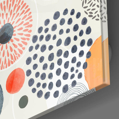 Close-up angled view of glass wall art showing central blue dot pattern surrounded by coral radiating lines, red oval, orange rectangle, and gray leaf designs on white background.