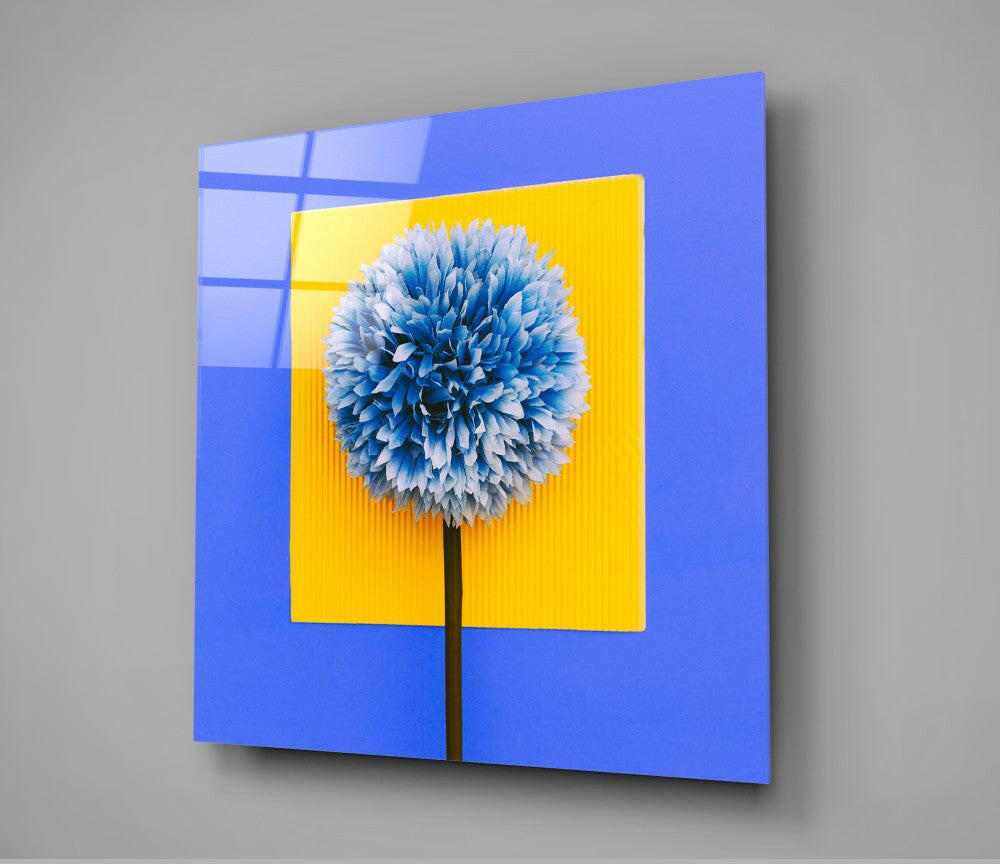 Blue glass wall art panel with yellow center frame displaying blue and white spherical flower against gray background, angled perspective view.