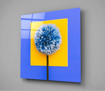 Blue glass wall art panel with yellow center frame displaying blue and white spherical flower against gray background, angled perspective view.